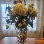 Floral arrangement in a vase on a wooden table with a window and curtain in the background