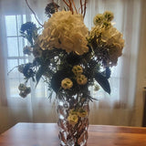 Floral arrangement in a vase on a wooden table with a window and curtain in the background